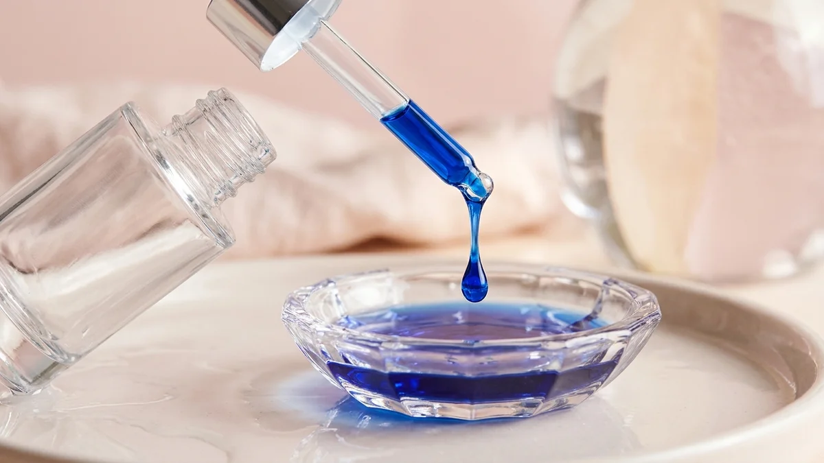 Frasco cuentagotas de cristal de lujo liberando una gota pura de sérum azul de péptido de cobre sobre un plato de cristal bajo luz suave de estudio